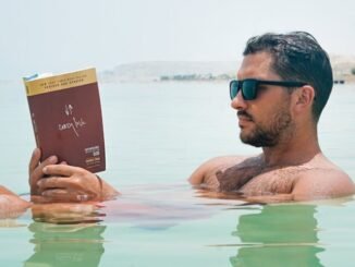 man wearing sunglasses reading book on body of water