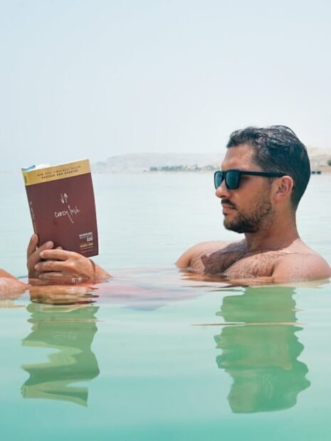 man wearing sunglasses reading book on body of water